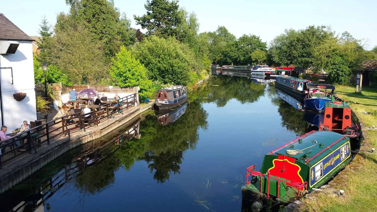 Moored narrowboats beside a canalside pub garden with trees reflected in the water
