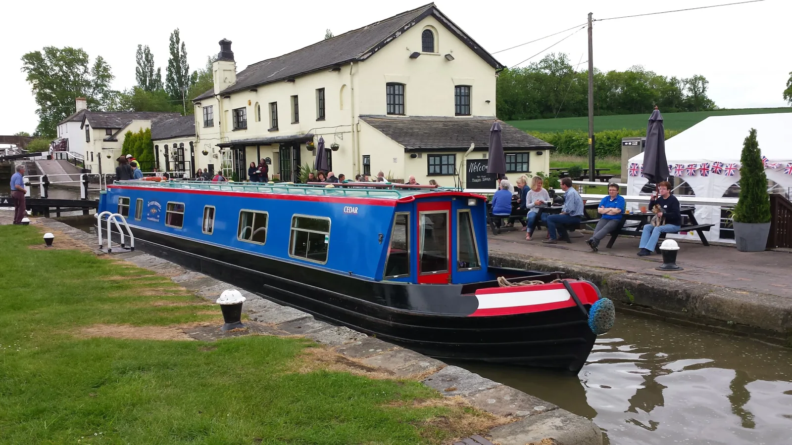 Blue narrowboat moored outside a canalside pub with people sitting at the tables