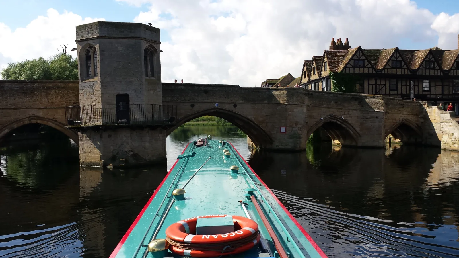 View from the bow of a boat approaching an old stone bridge with a tower beside it