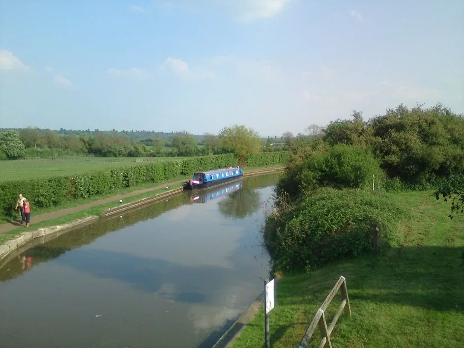 Narrowboat cruising a straight stretch of canal between grassy banks and hedgerows