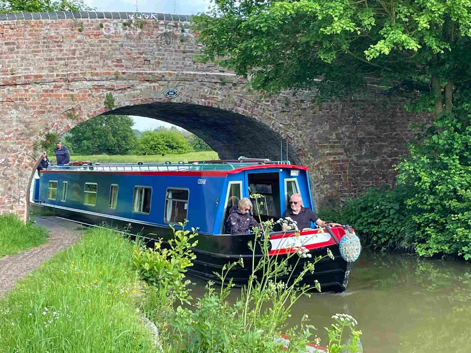Blue narrowboat passing under a brick arch bridge with two people on the stern