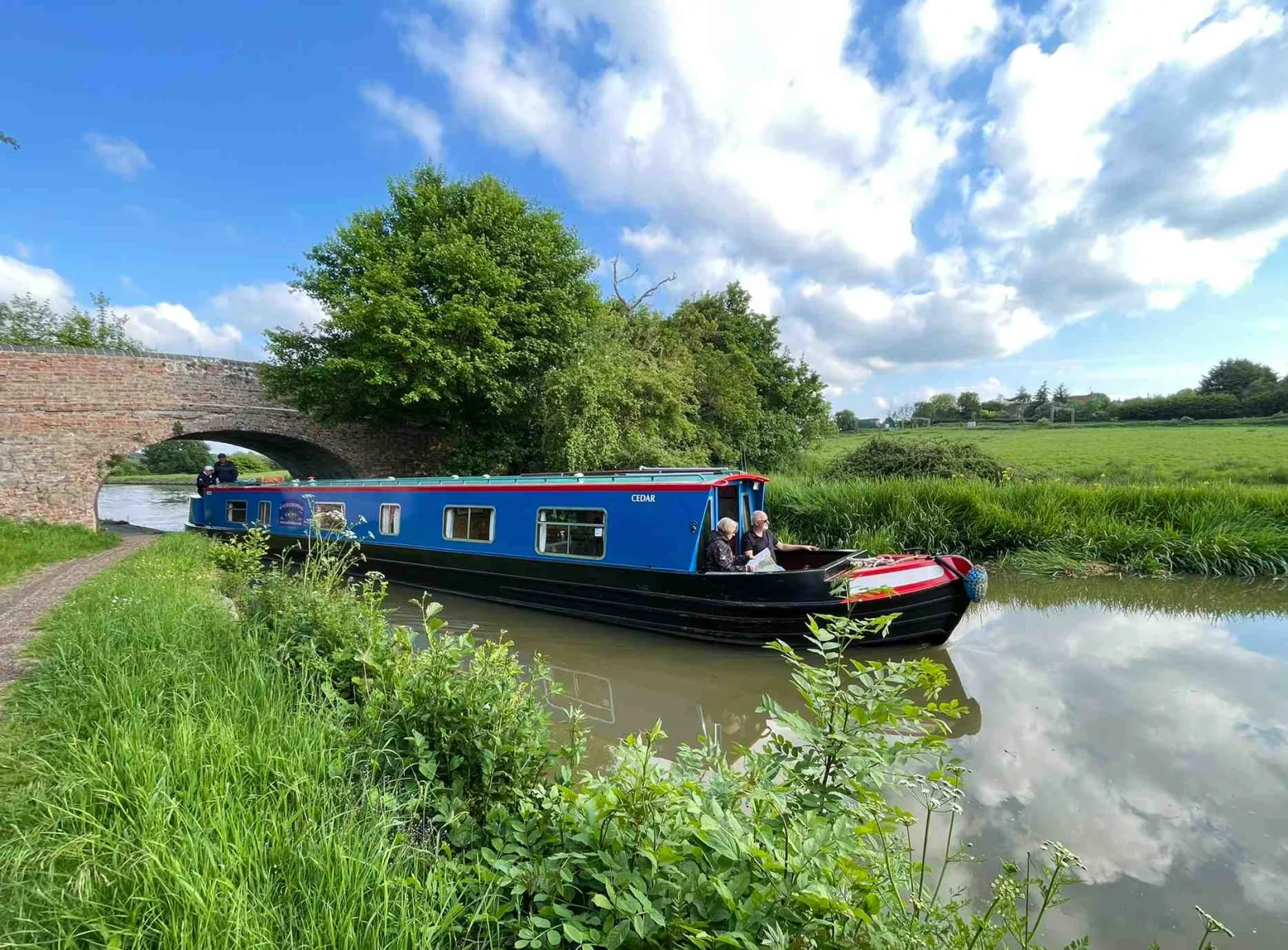 Blue narrowboat cruising past open fields under a blue sky