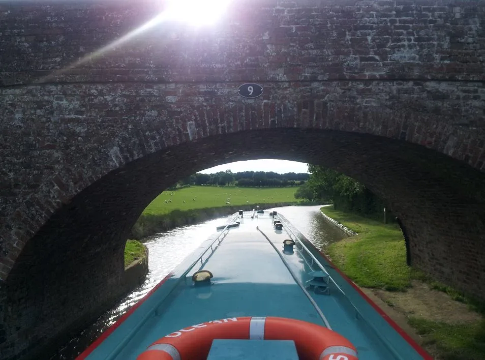 View from the bow of a narrowboat entering a brick bridge hole with the sun above