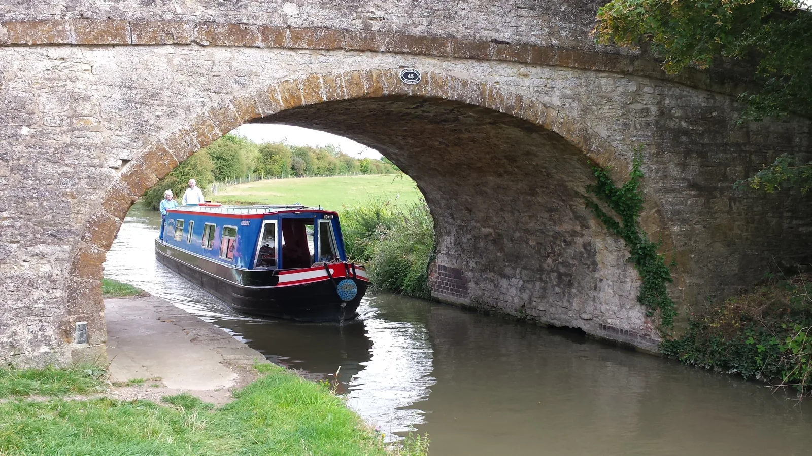 Blue narrowboat passing under a stone hump-backed bridge on a quiet canal