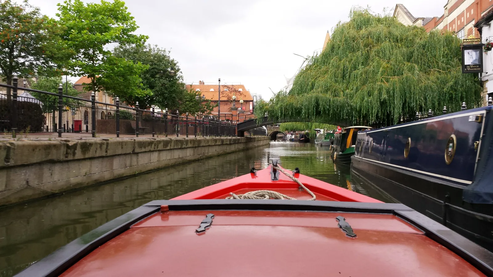 View over the bow of a narrowboat passing moored boats beside a high town wall