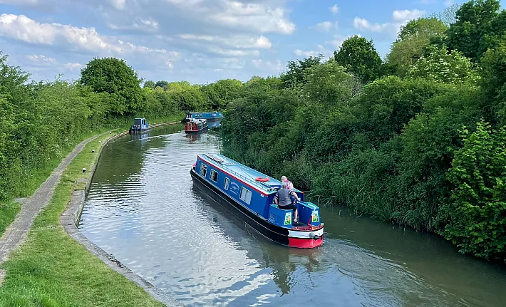 Blue narrowboat cruising a tree-lined stretch of canal with more boats moored ahead
