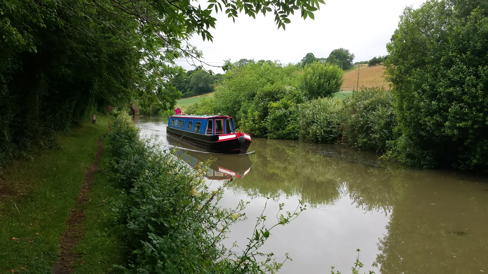 Narrowboat cruising a quiet canal through open countryside with trees along the bank