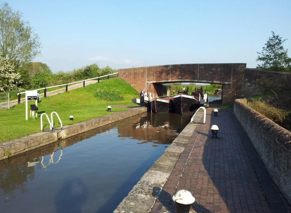 Narrowboats moored below a brick bridge with lock gates and a walkway in the foreground
