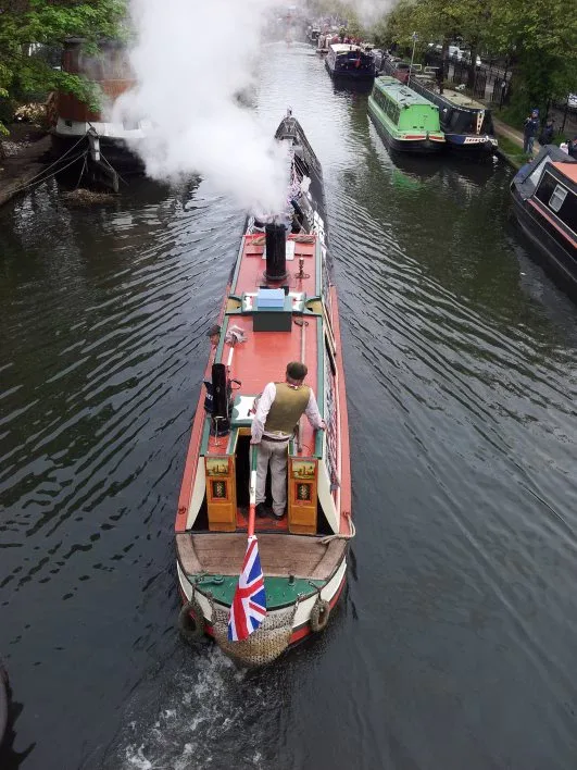 Traditional narrowboat with a smoking chimney cruising past moored boats and flying a Union flag