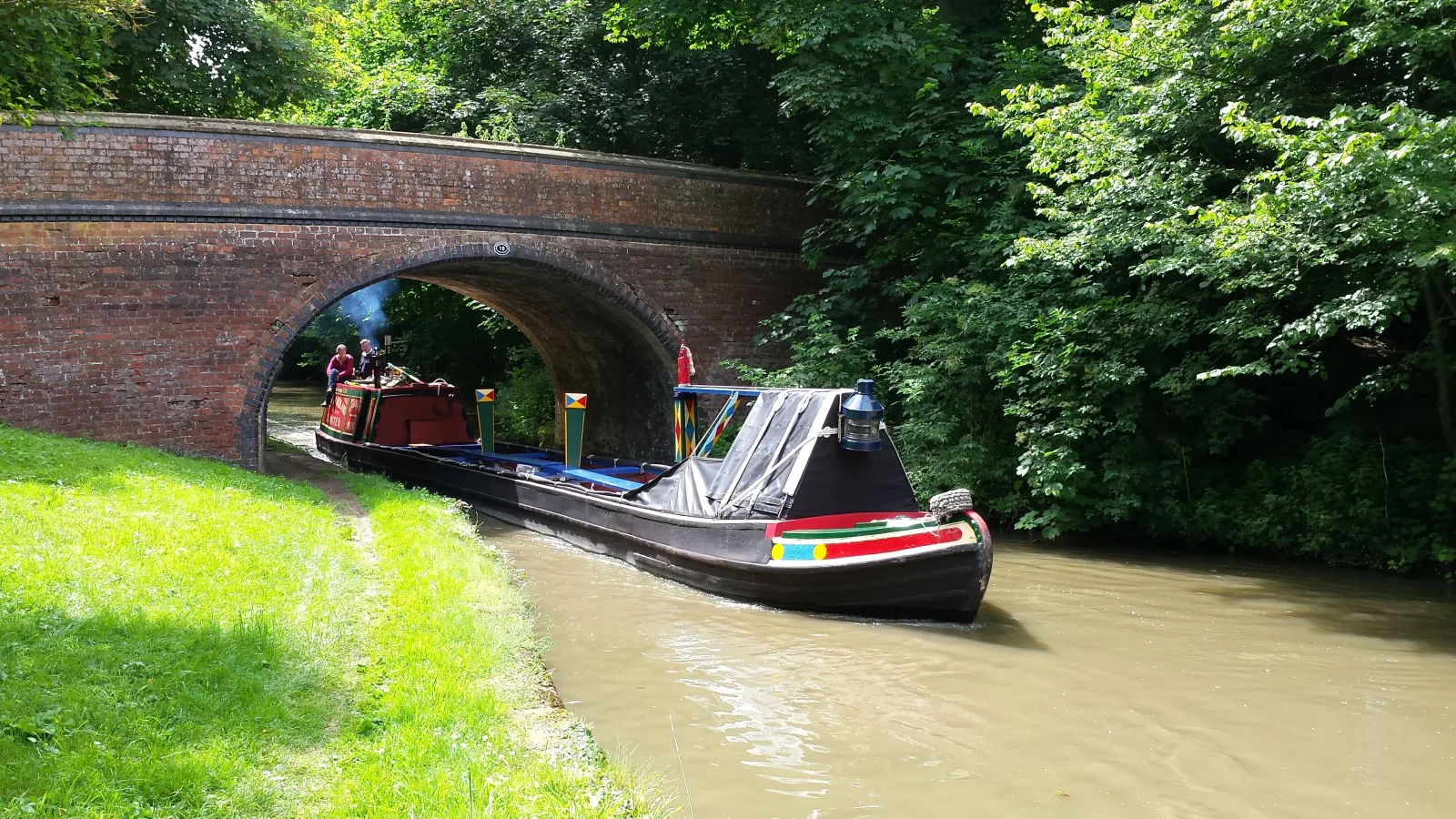 Dark green narrowboat passing under a red brick bridge on a sunny day