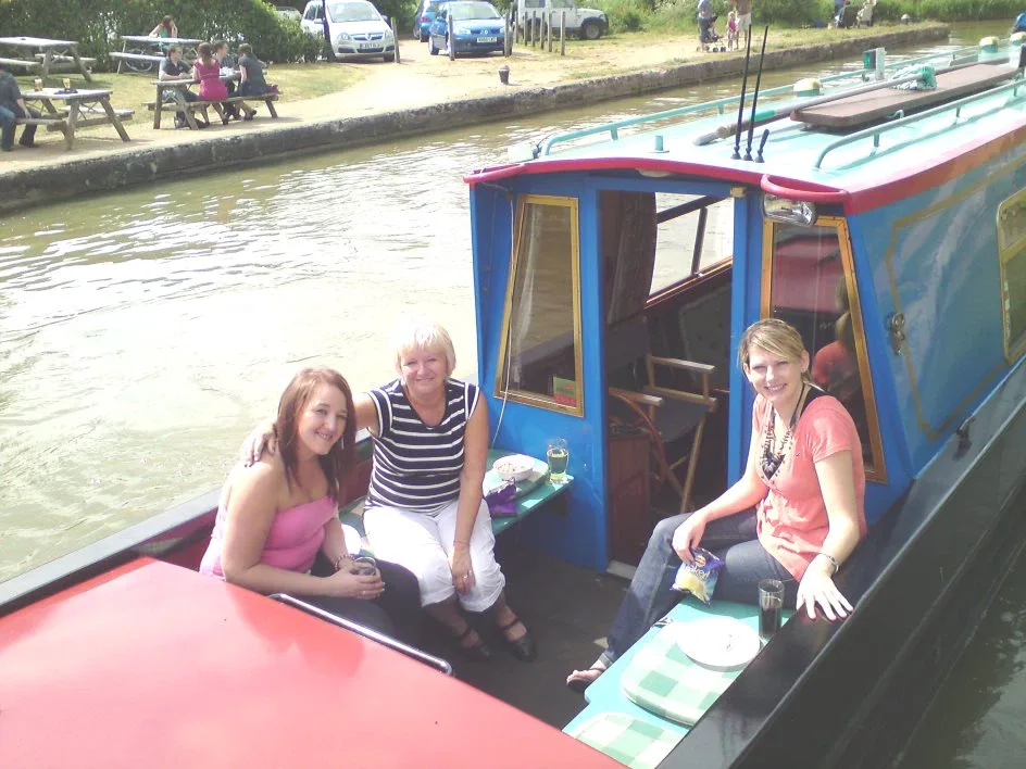 Three women sitting on the stern of a blue narrowboat moored on the towpath