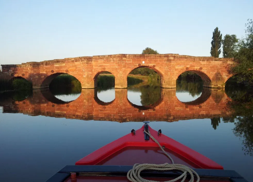 Wide stone bridge with five arches reflected in still water seen from a boat