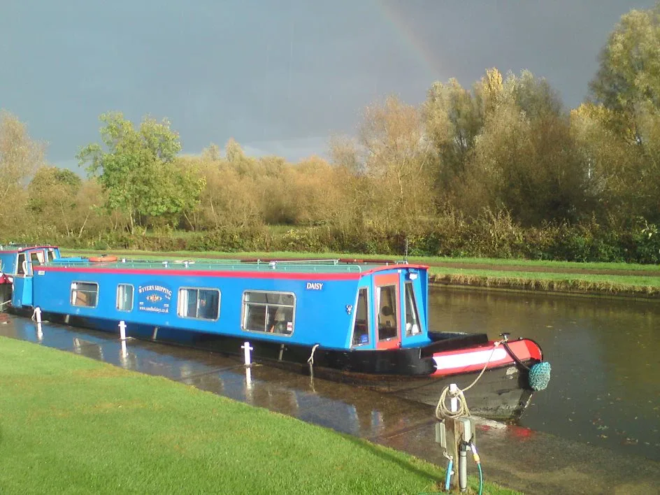 Blue narrowboat cruising past autumn trees with a rainbow in the sky behind