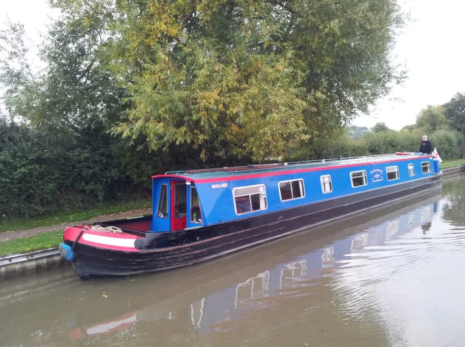 Blue narrowboat cruising past a wooded bank on a still stretch of canal