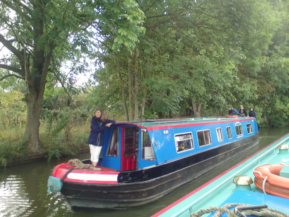 Blue narrowboat moored under a large tree with a crew member standing on the bow